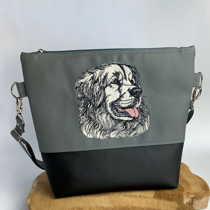 Umhängetasche aus Kunstleder, bestickt mit einer Applikation aus hellem Kunstfell, das Stickmotiv von einem Hovawart-Kopf wird in schwarz darüber gestickt. die Tasche ist oben dunkelgrau und unten schwarz.