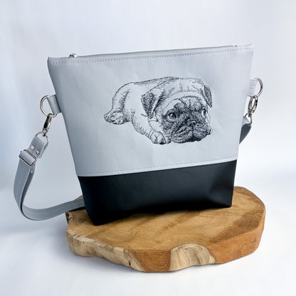 Umhängetasche/Handtasche aus Kunstleder mit Mops-Motiv bestickt, zweifarbig, oben hellgrau, unten schwarz. der Mops ist in schwarzer Kontur detailreich gestickt. liegt auf den Vorderpfoten